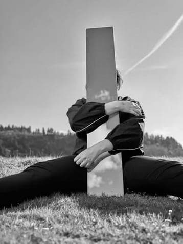 Person sitting on grass and hugging a tall mirror that reflects the clouds